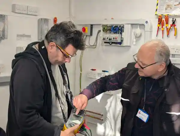 Sezai Aramaz demonstrating the use of a multifunction tester to a trainee electrician during a practical session in a training workshop, with tools, wiring, and electrical boards visible in the background.